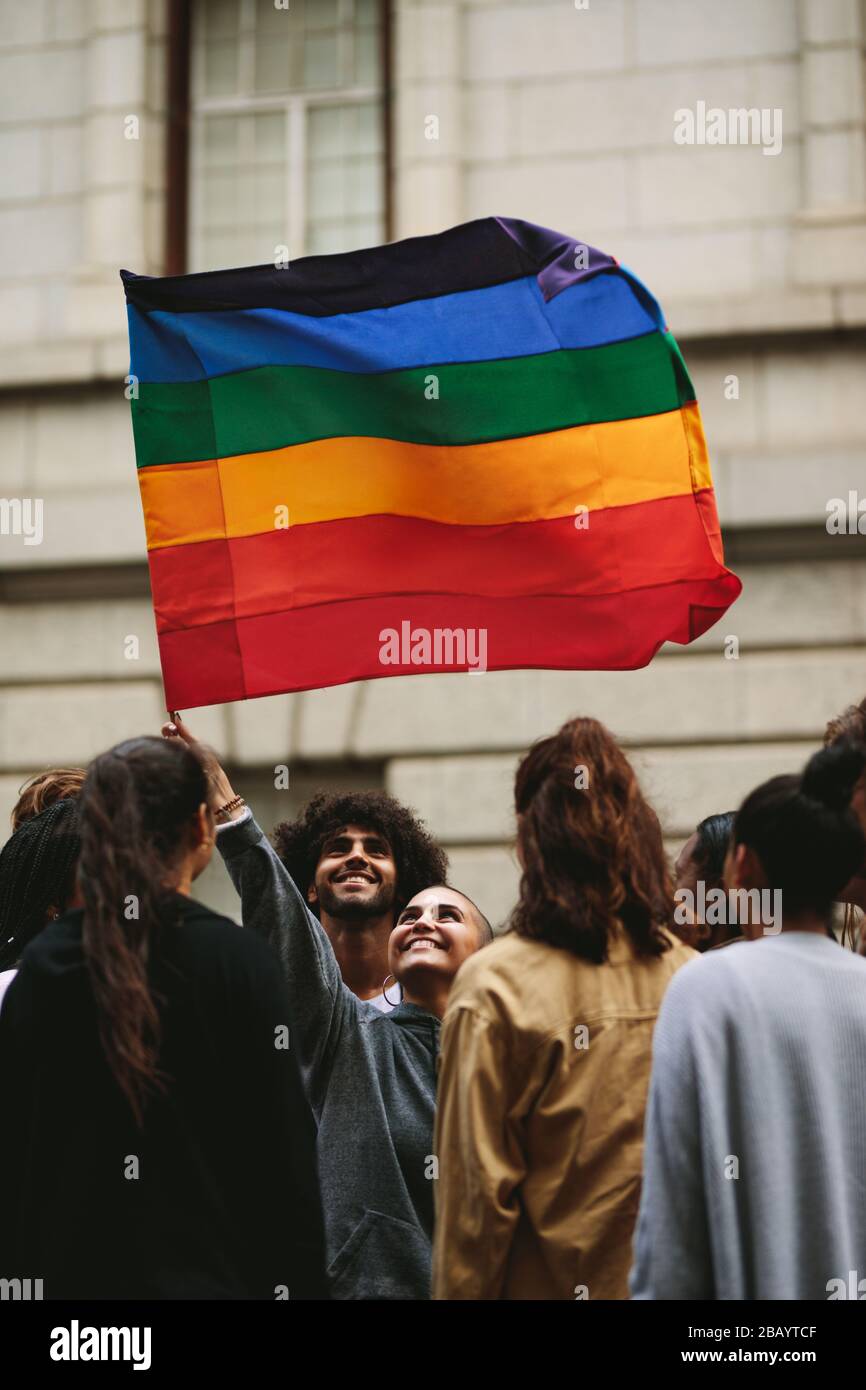 Smiling woman waving gay flag with group of people standing around. People participating in Gay march in the city. Stock Photo