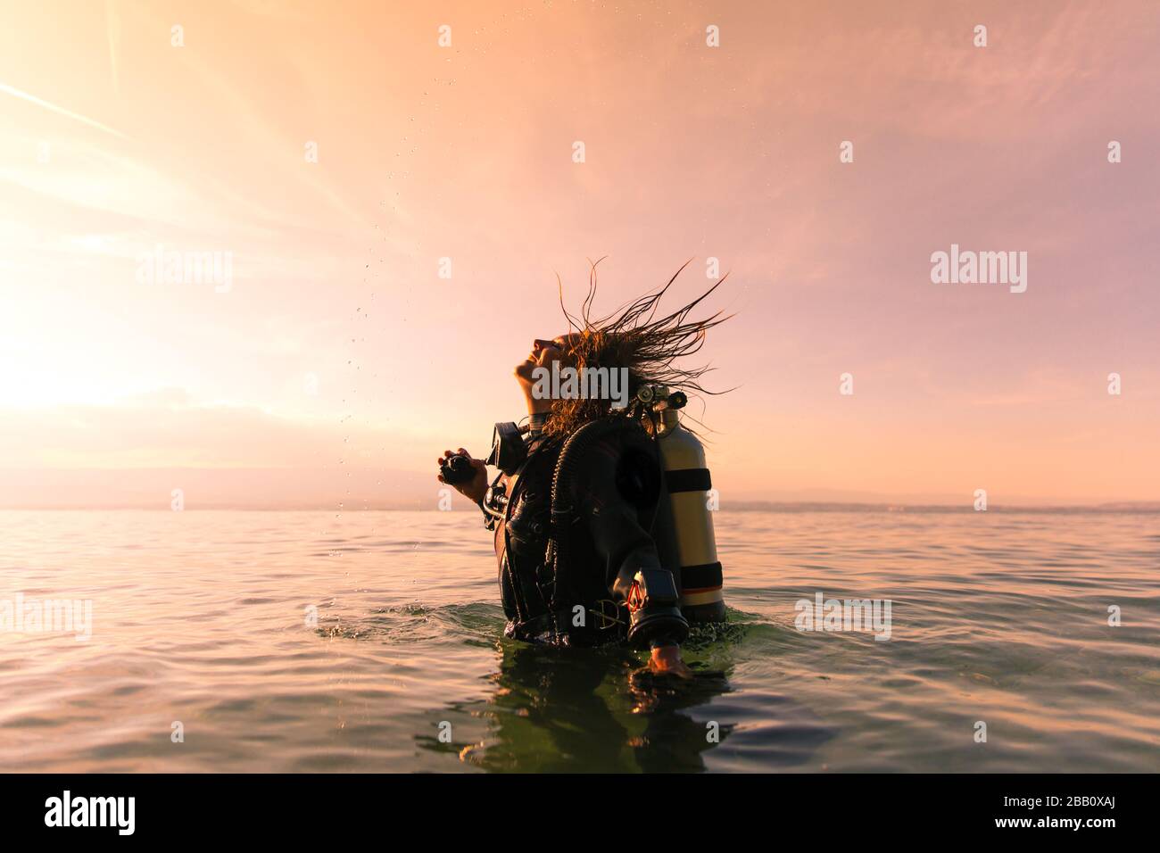 Female Scuba Dive Instructor Wearing a Dry Suit, a Twin Tank and Holding Fins Flipping Wet Hair in the Air Stock Photo