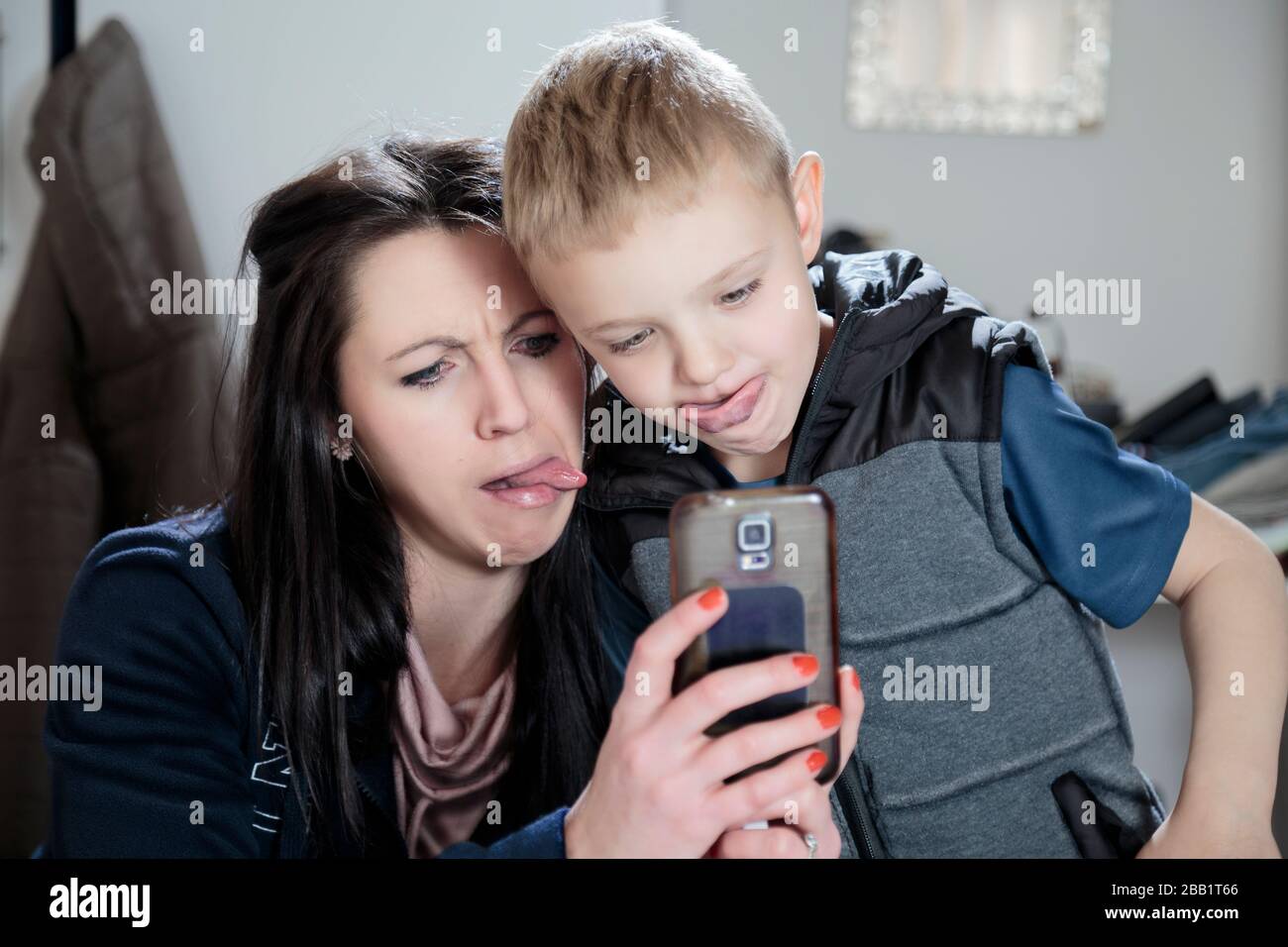 A color photo of a beautiful young mother and her boy looking at a mobile phone and pulling funny faces with they're tongues out. Stock Photo