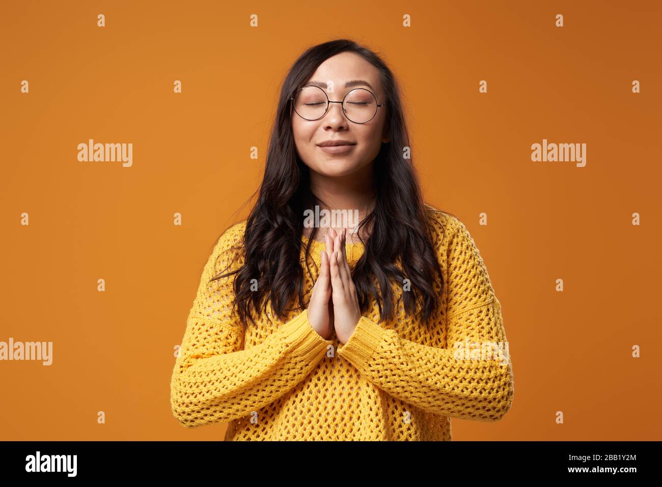 Praying young asian woman with closed eyes wearing glasses and in yellow knitted sweater on empty orange background in studio Stock Photo