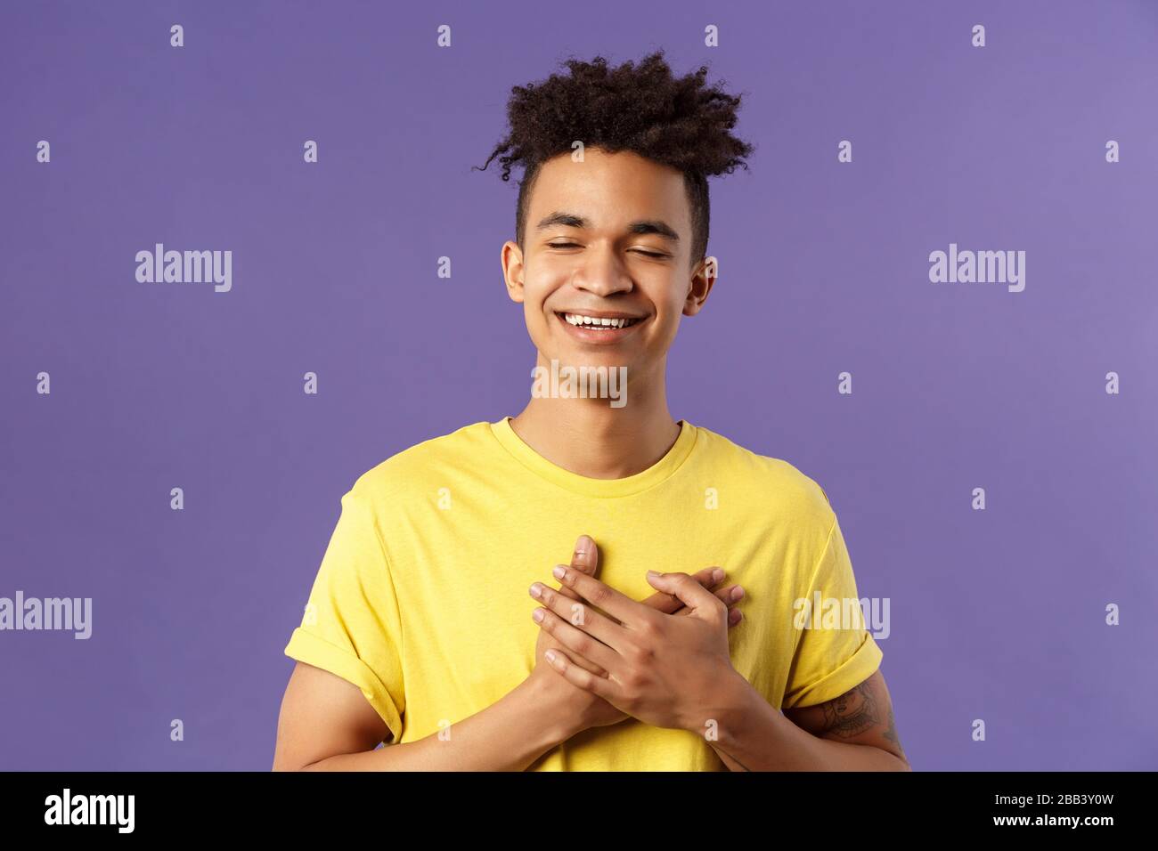 Close-up portrait of happy, upbeat young dreamy guy, remember sweet memories, hold hands on heart, smiling touched and delighted, close eyes grinning Stock Photo