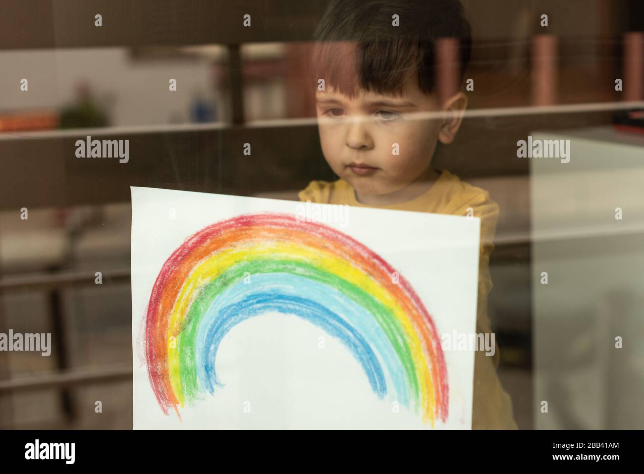 Inspirational little kid holding a drawing of a rainbow through the window Stock Photo