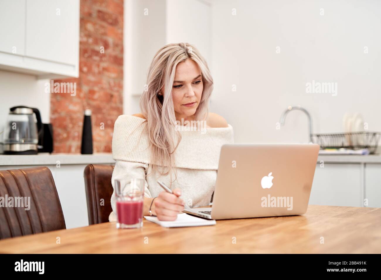 A single caucasian female sits at a table in the kitchen whilst working on a laptop with a healthy juice drink next to her in a glass Stock Photo