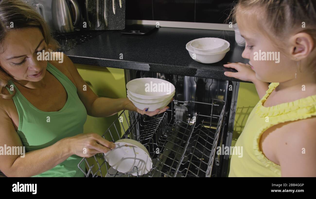 Mother teaching daughter smart girl learning to use dishwasher. Young