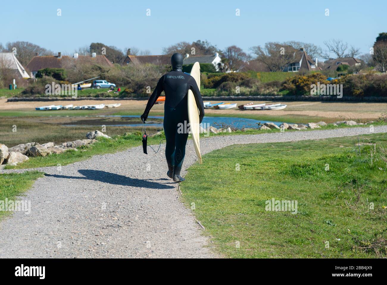 Surfer in full wetsuit walks along footpath to village in March after surfing in the cold water on West Wittering Beach, Chichester,West Sussex, UK Stock Photo