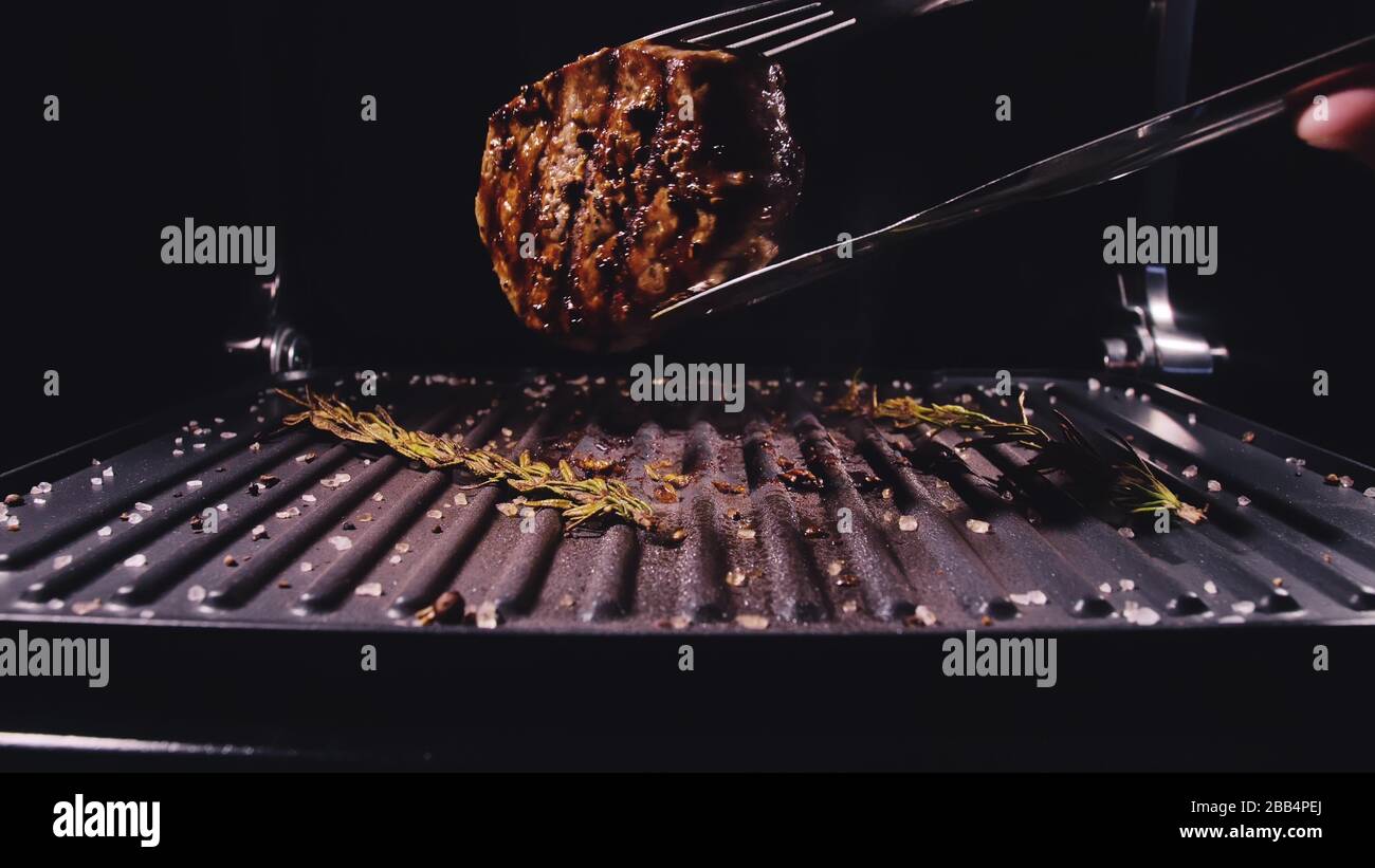 Delicious juicy meat steak cooking on grill. Aged prime rare roast grilling marble beef. Electric roaster, rosemary black pepper, salt. Kitchen tongs. Stock Photo