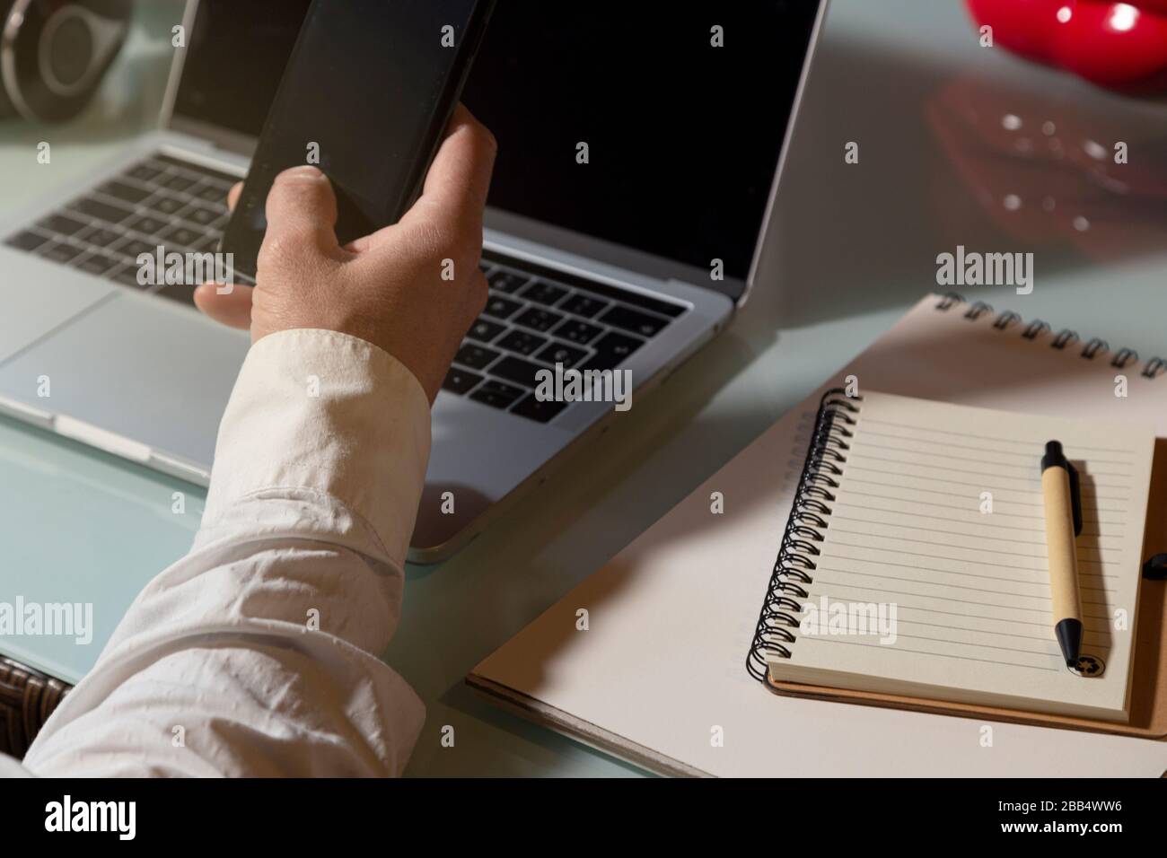 The hand of a man who uses the mobile phone and laptop to work from home. Note pad and pen on the table. Telework concept Stock Photo