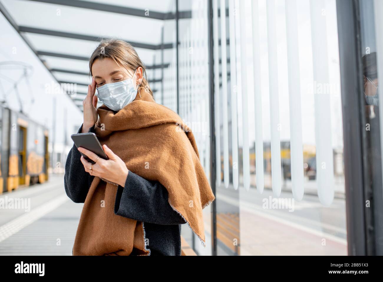 Young and depressed woman in face mask worried reading bad news on a smart phone at a public transport stop during an epidemic. Concept of panic and bad influence of news Stock Photo