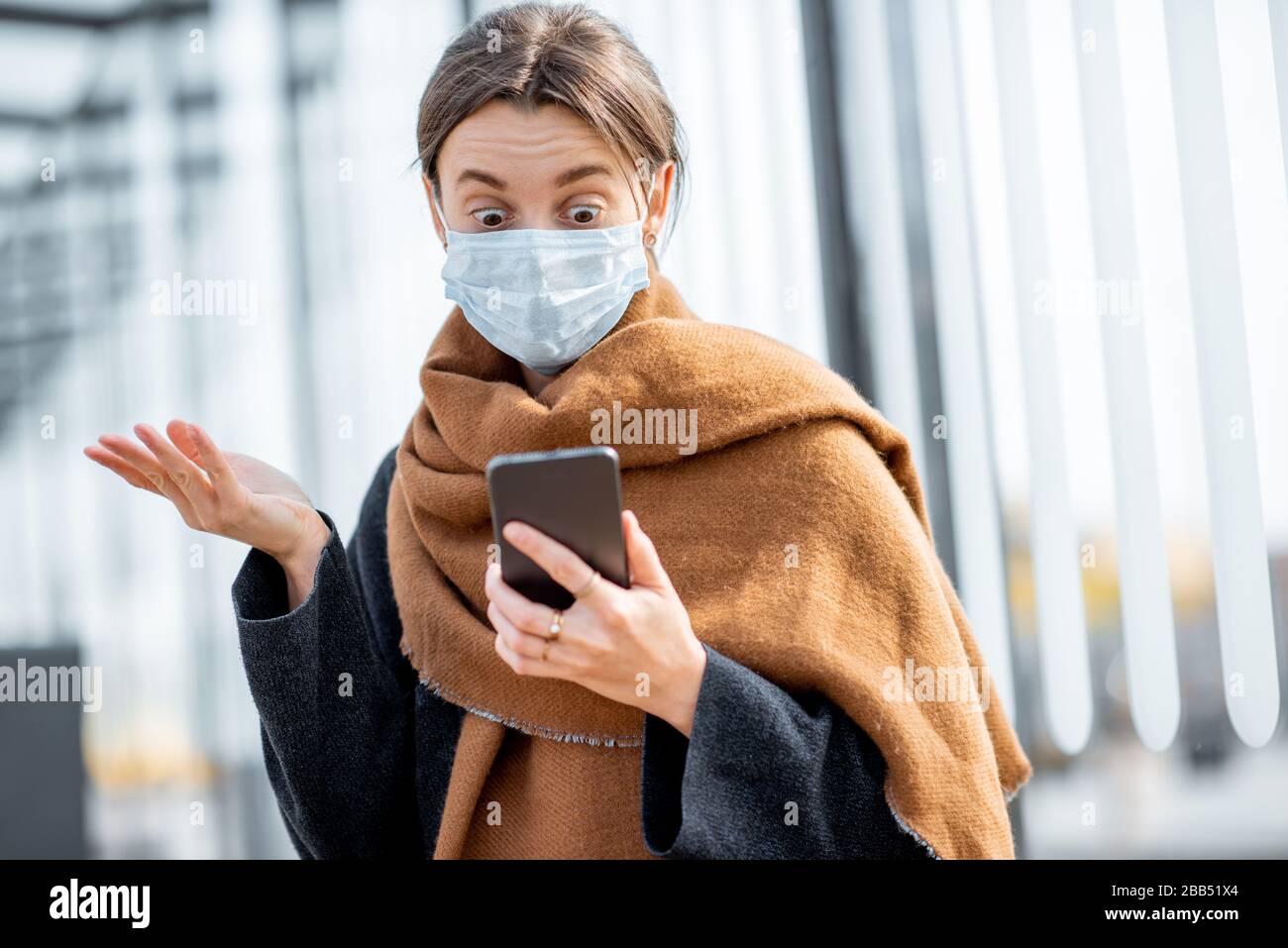 Young and depressed woman in face mask worried reading bad news on a smart phone at a public transport stop during an epidemic. Concept of panic and bad influence of news Stock Photo