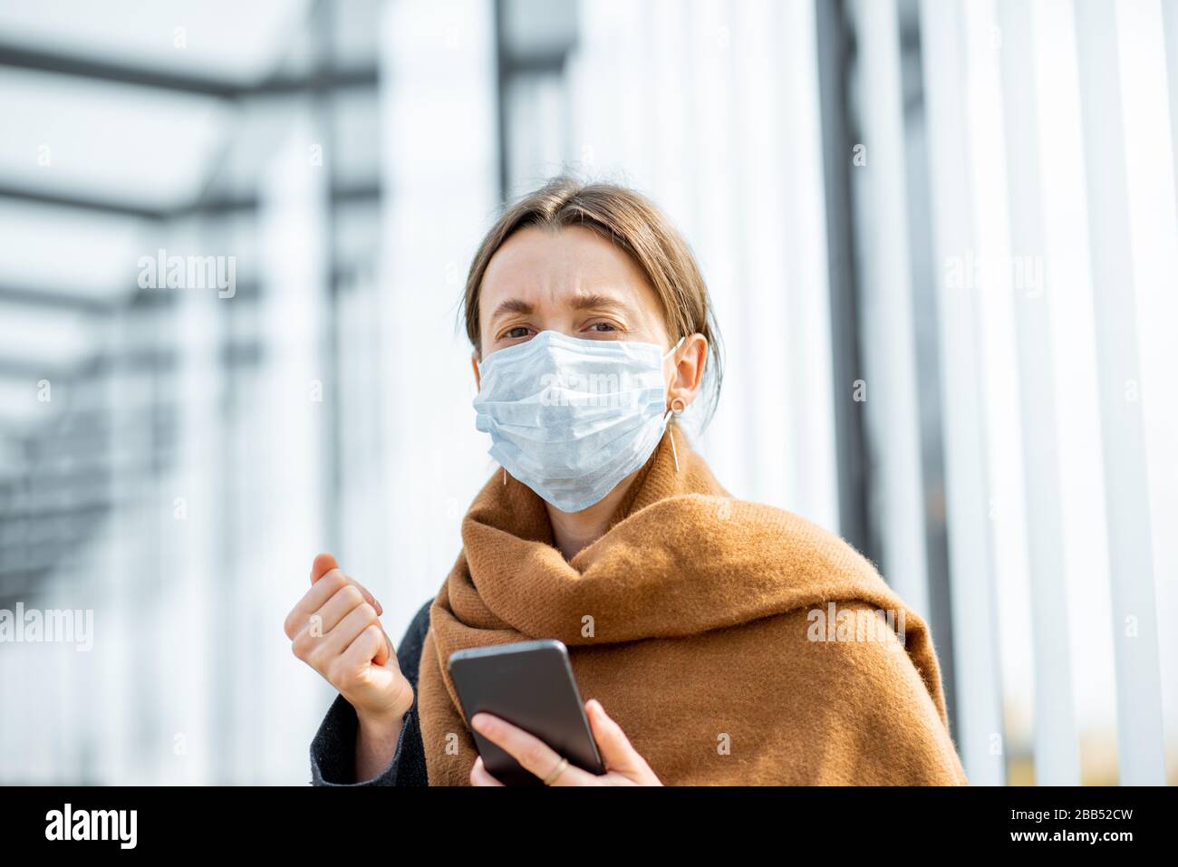 Young and depressed woman in face mask worried reading bad news on a smart phone at a public transport stop during an epidemic. Concept of panic and bad influence of news Stock Photo