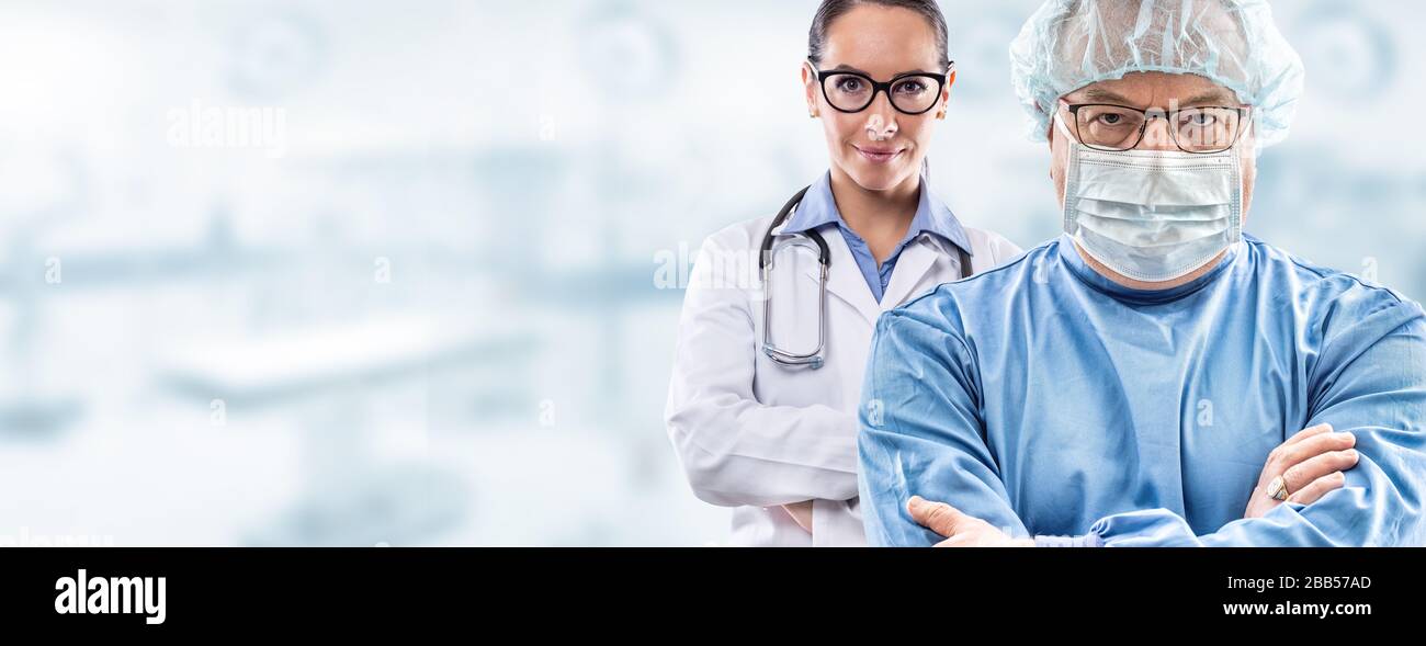 Male mid age surgeon in glasses wearing blue dress, protective face mask and hat; young female doctor in white dress with glasses and stethoscope stan Stock Photo