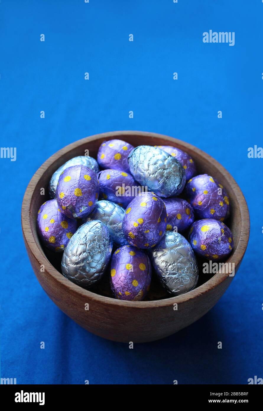 A wooden bowl filled with colorful chocolate Easter eggs in foil wrappings, with a blue table cloth background. High angle view and copy space above. Stock Photo
