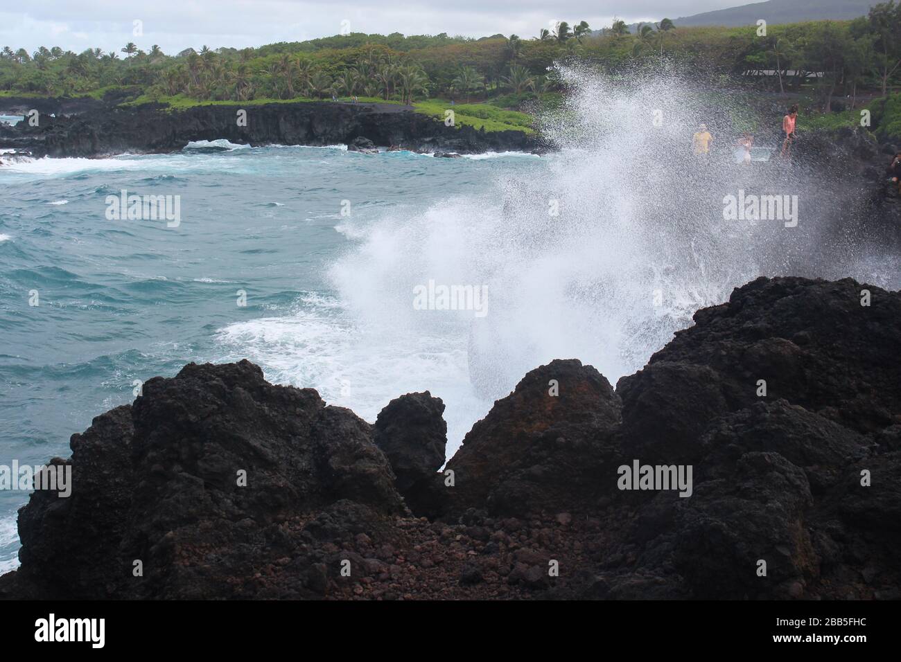 The sea spray from a wave in the Pacific Ocean crashing into volcanic rocks and soacking tourists at Waianapanapa State Park in Hana, Maui, Hawaii, US Stock Photo