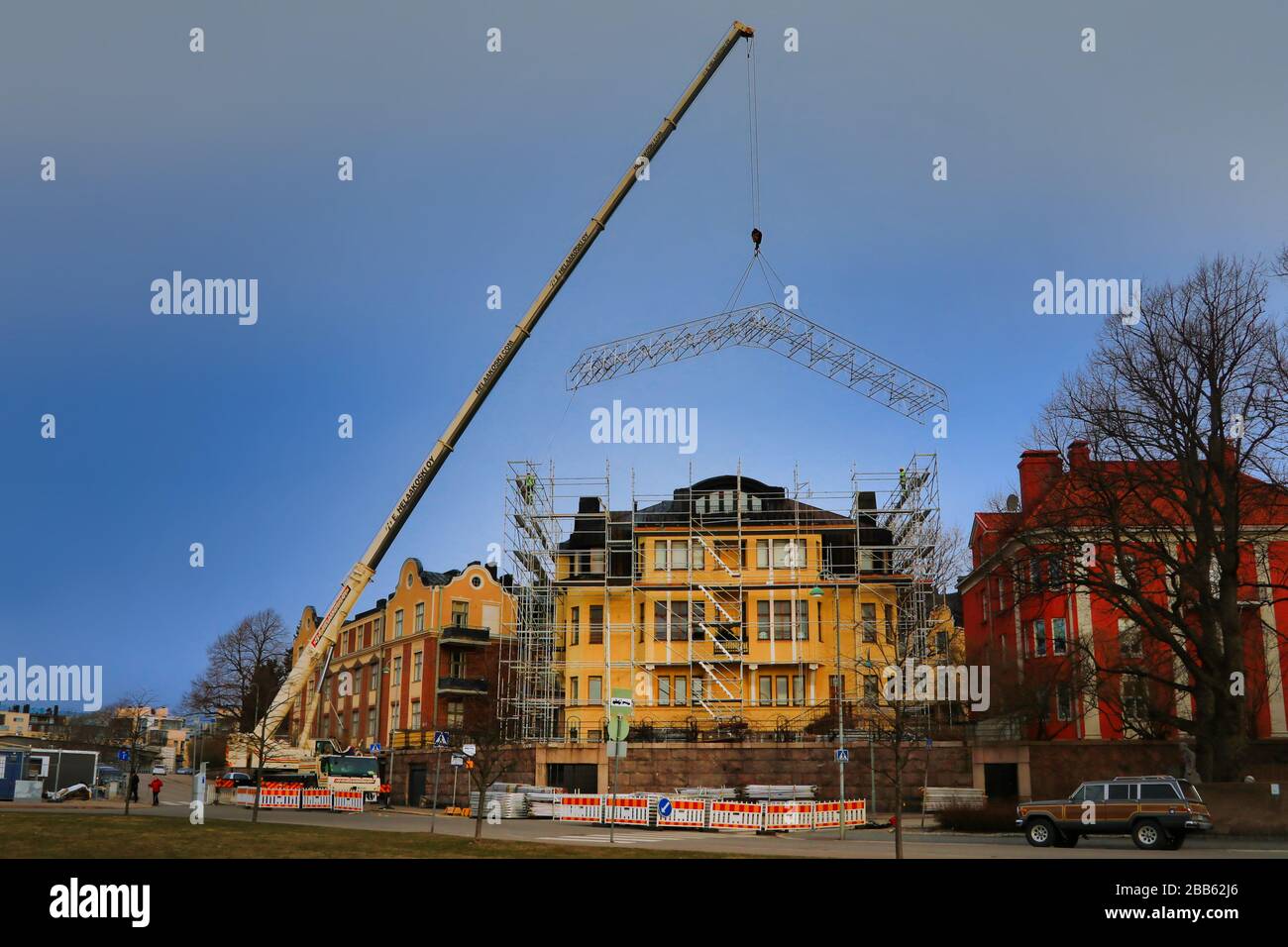 Mobile crane truck E. Helaakoski Oy is lifting a large construction metal frame to the roof top in Helsinki, Finland. March 30, 2020. Stock Photo