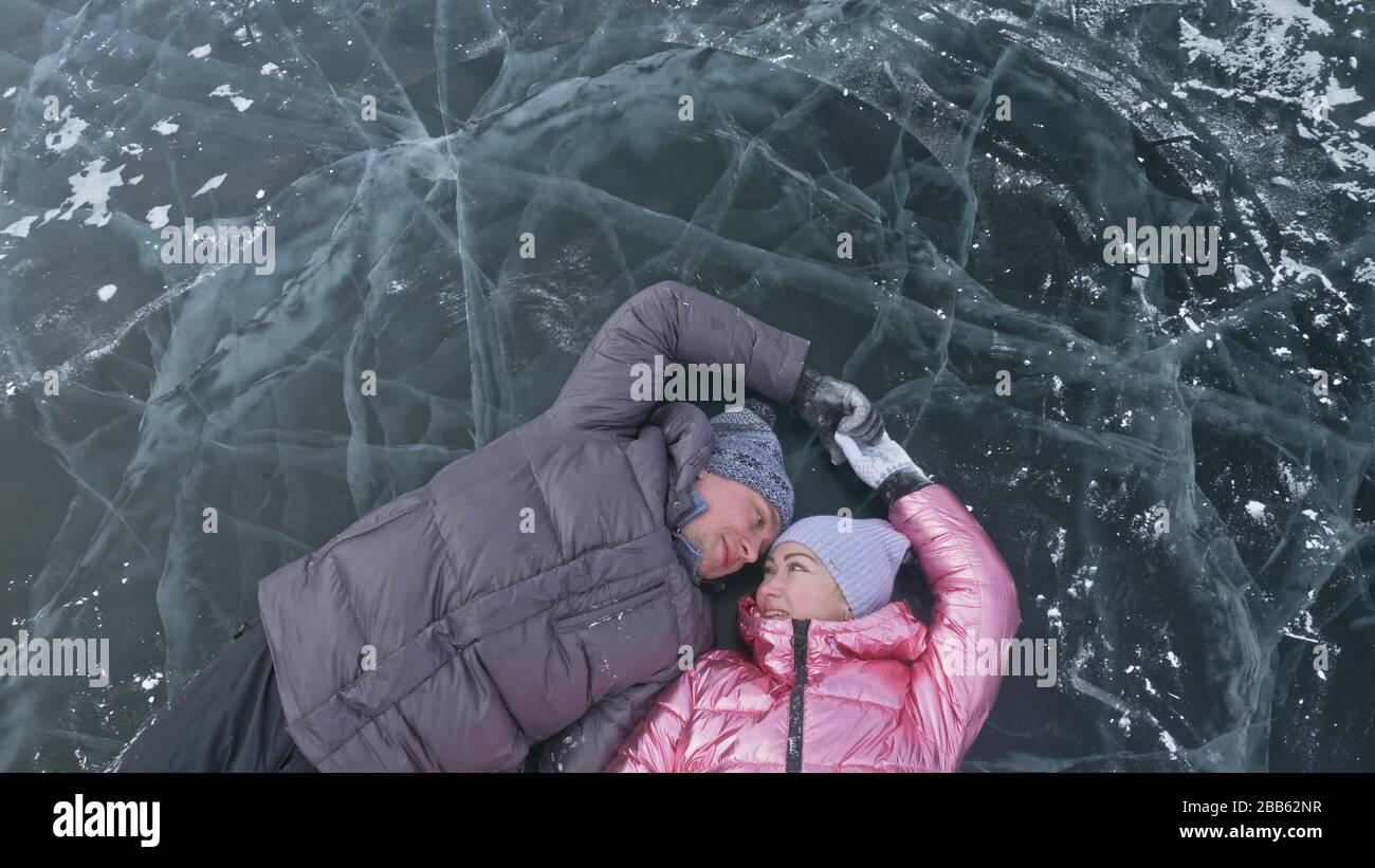 Couple has fun during winter walk against background of ice of frozen lake. Lovers lie on clear ice, have fun, kiss and hug. View from above. Stock Photo