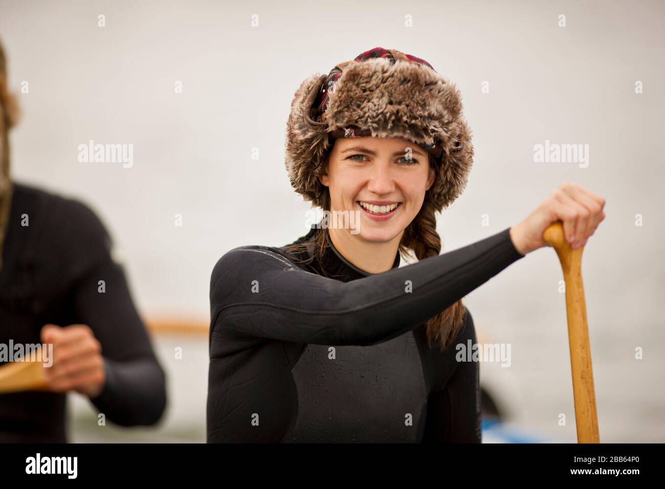 Portrait of a young woman wearing a fur hat and wetsuit while paddling in canoe with friends. Stock Photo
