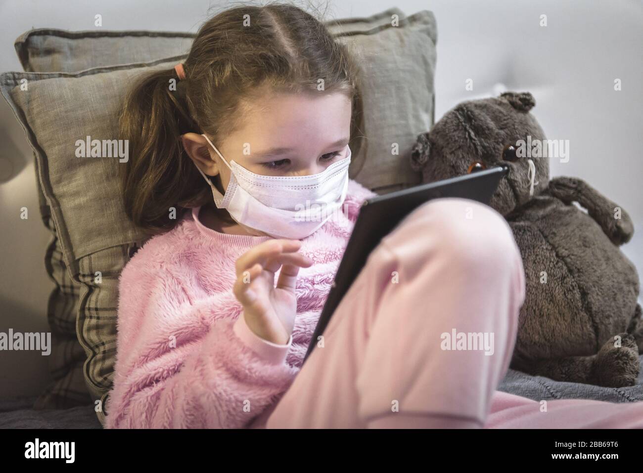 COVID-19 coronavirus quarantine concept, little girl in medical mask plays with digital tablet at home. Kid uses computer in room during COVID coronav Stock Photo