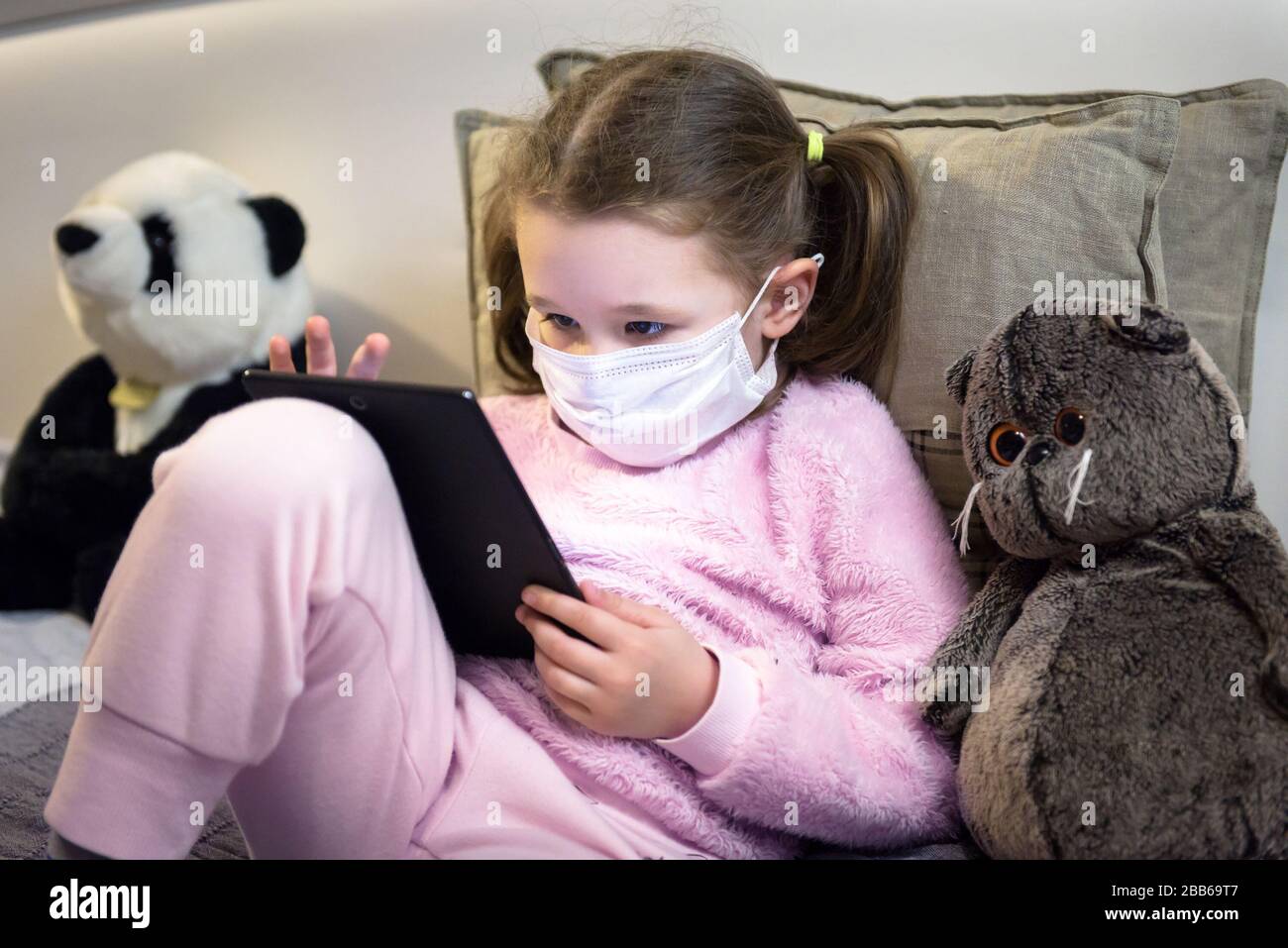 COVID-19 coronavirus quarantine concept, little girl in face mask plays with digital tablet at home. Kid using computer in room during COVID coronavir Stock Photo