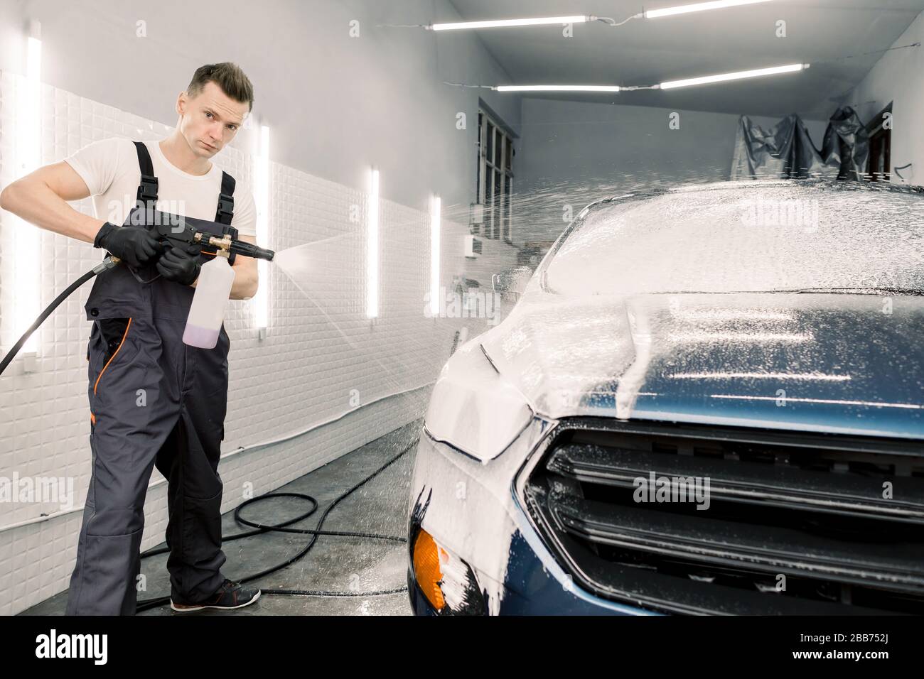 A man holds a foam sprayer from which foam flies out onto a car. A car at a  self service car wash. Stock Photo by romanchoknadii