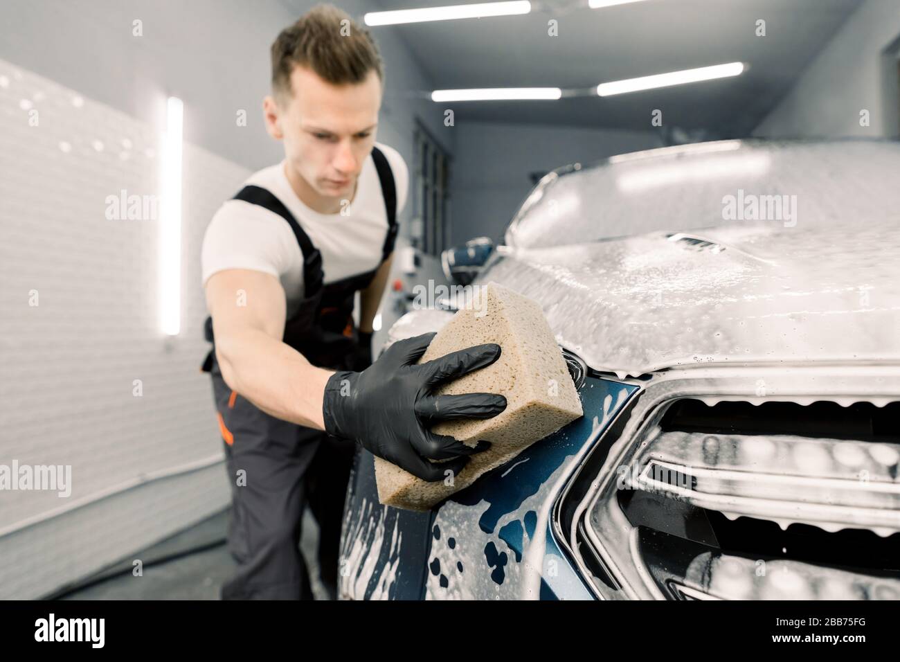 Young man car wash worker washing a soapy dark blue car, holding sponge