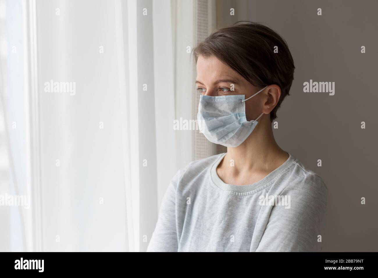 A woman in a medical protective mask is sad at the window. Stock Photo