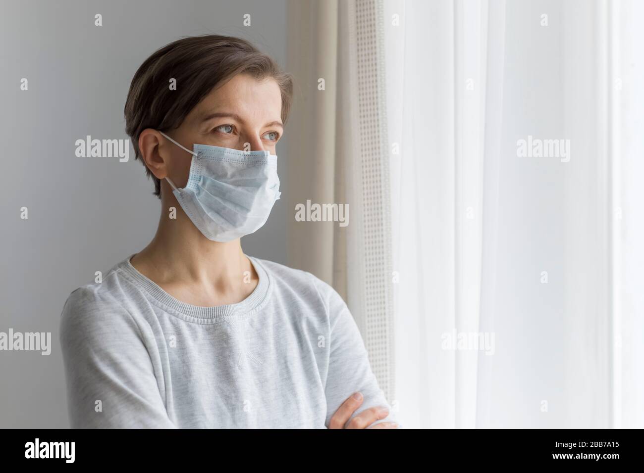 A woman in a medical protective mask is sad at the window. Stock Photo