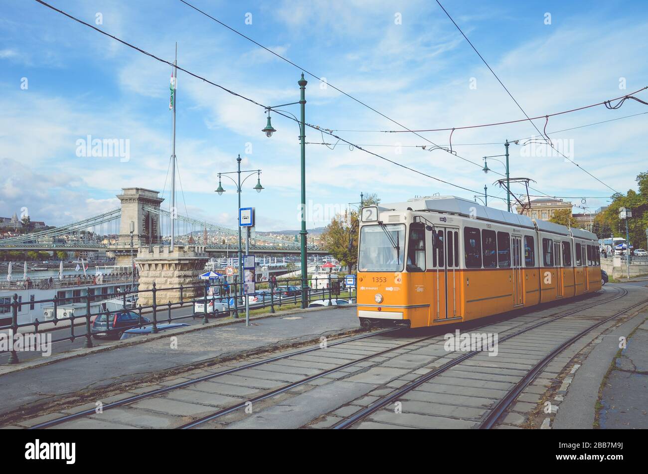 Budapest, Hungary - Nov 6, 2019: Yellow tram at a tram station. Szechenyi chain bridge and the historical center in the background. Hungarian public transport. Horizontal photo with filter. Stock Photo