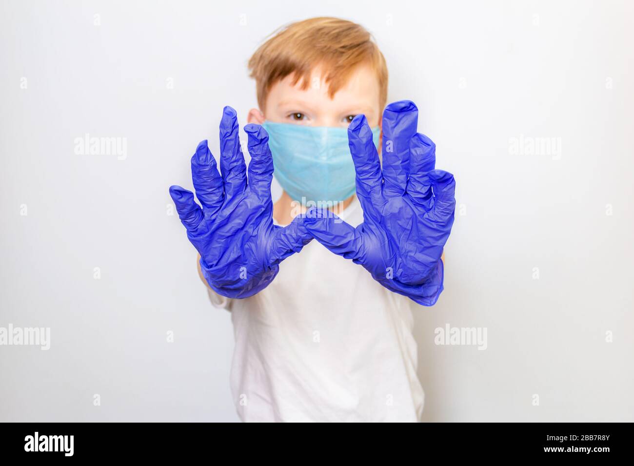 A seven-year-old boy in a medical mask and latex gloves extended his arms in front of him on a white background. Social distance, caronavirus Stock Photo