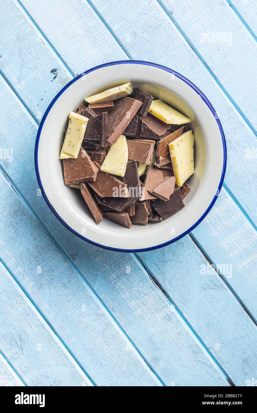 Broken white, milk and dark chocolate bars in bowl on blue wooden table. Stock Photo
