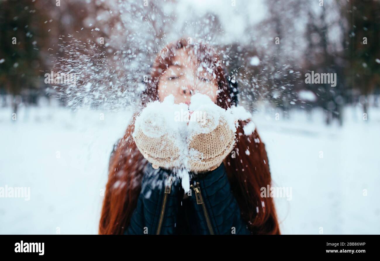 Winter. Woman with red hair wearing ear muffs blowing on snow in hands Stock Photo