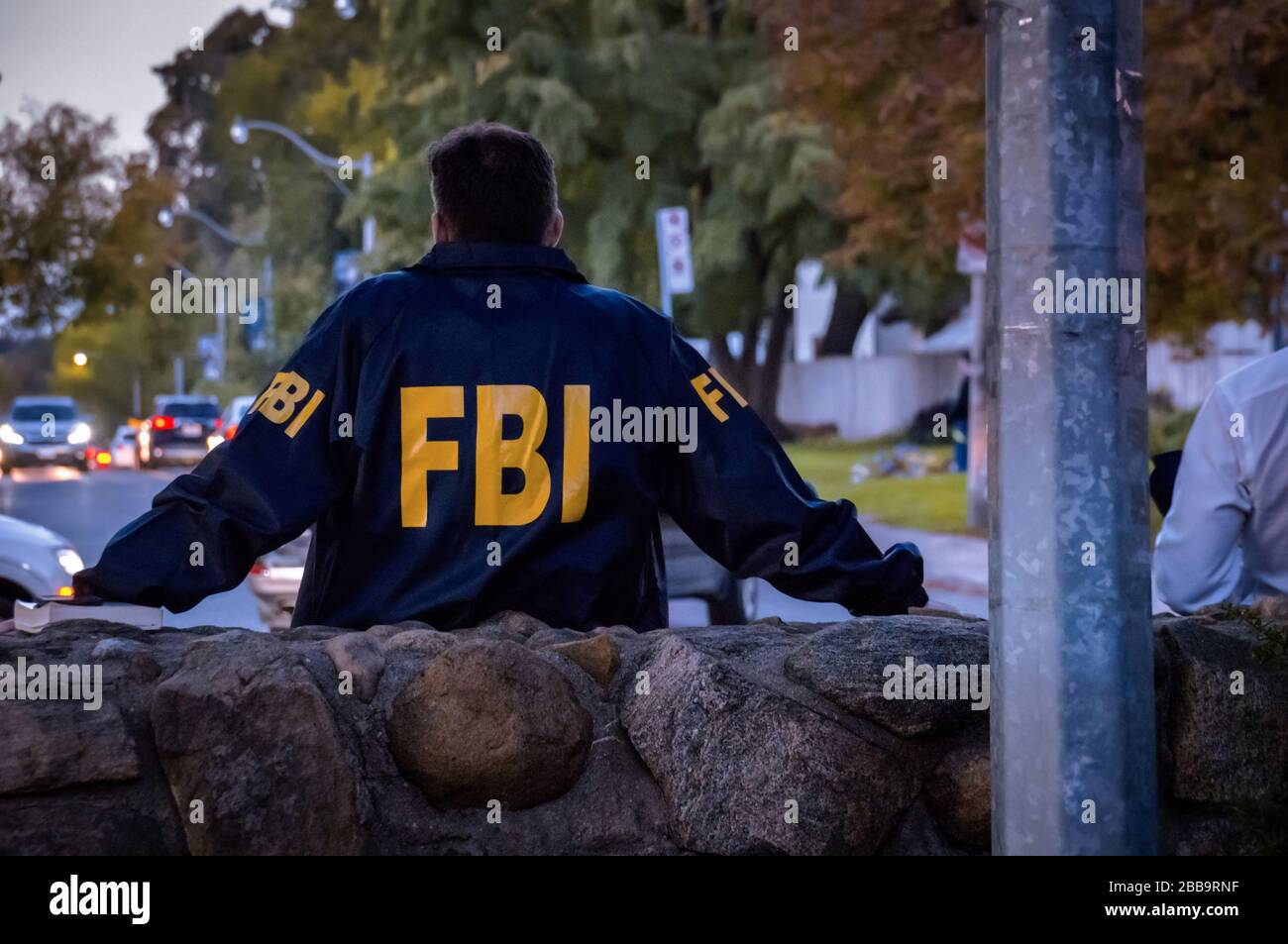 Male FBI agent wearing dark blue coat with FBI logo looking down the street with cars in the dusk seen from behind Stock Photo