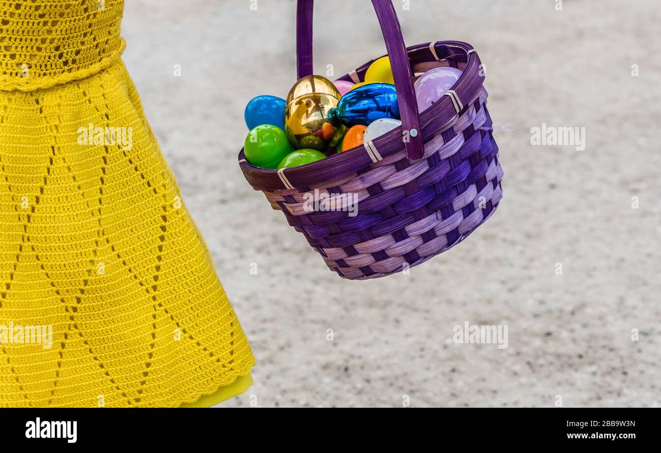 A purple easter egg basket with shiny colored plastic easter eggs in it being held by a child in a yellow dress. Stock Photo