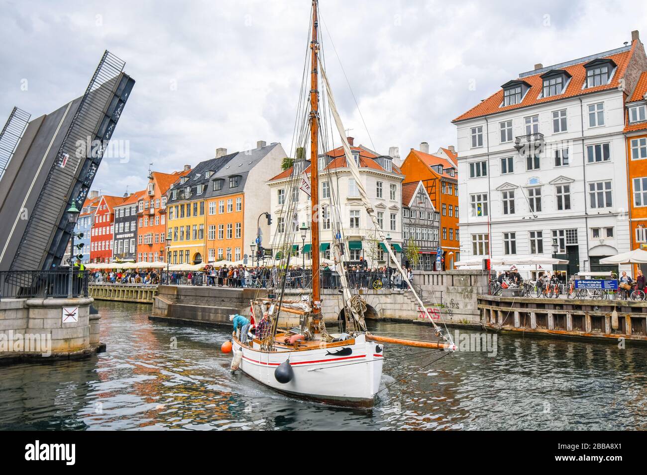 A sailboat glides through and under the opened bridge on the Nyhavn Canal in Copenhagen Denmark as tourists watch from shops and cafes alongside. Stock Photo