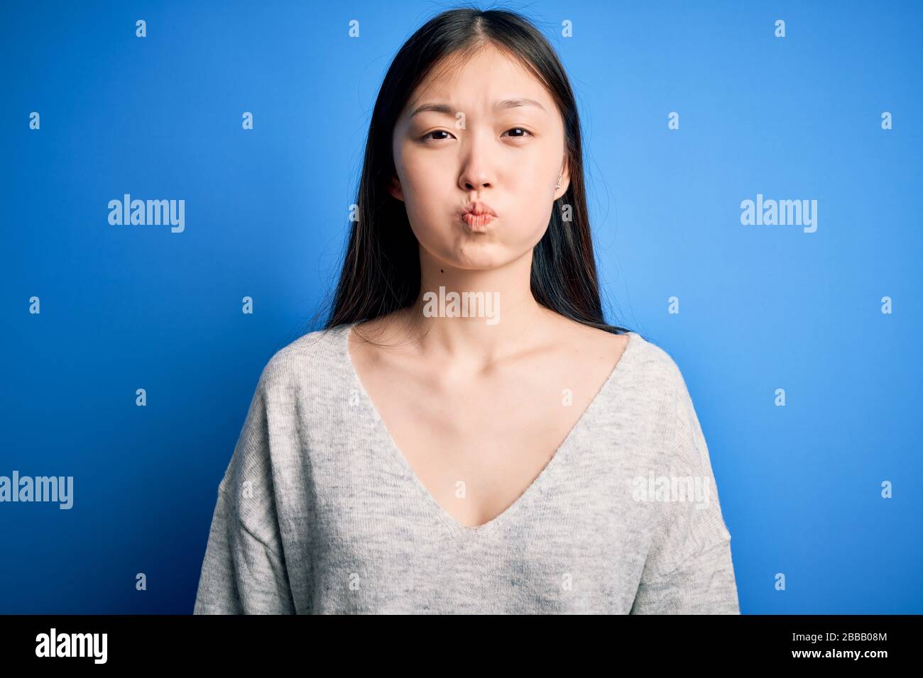 Young beautiful asian woman wearing casual sweater standing over blue isolated background puffing cheeks with funny face. Mouth inflated with air, cra Stock Photo