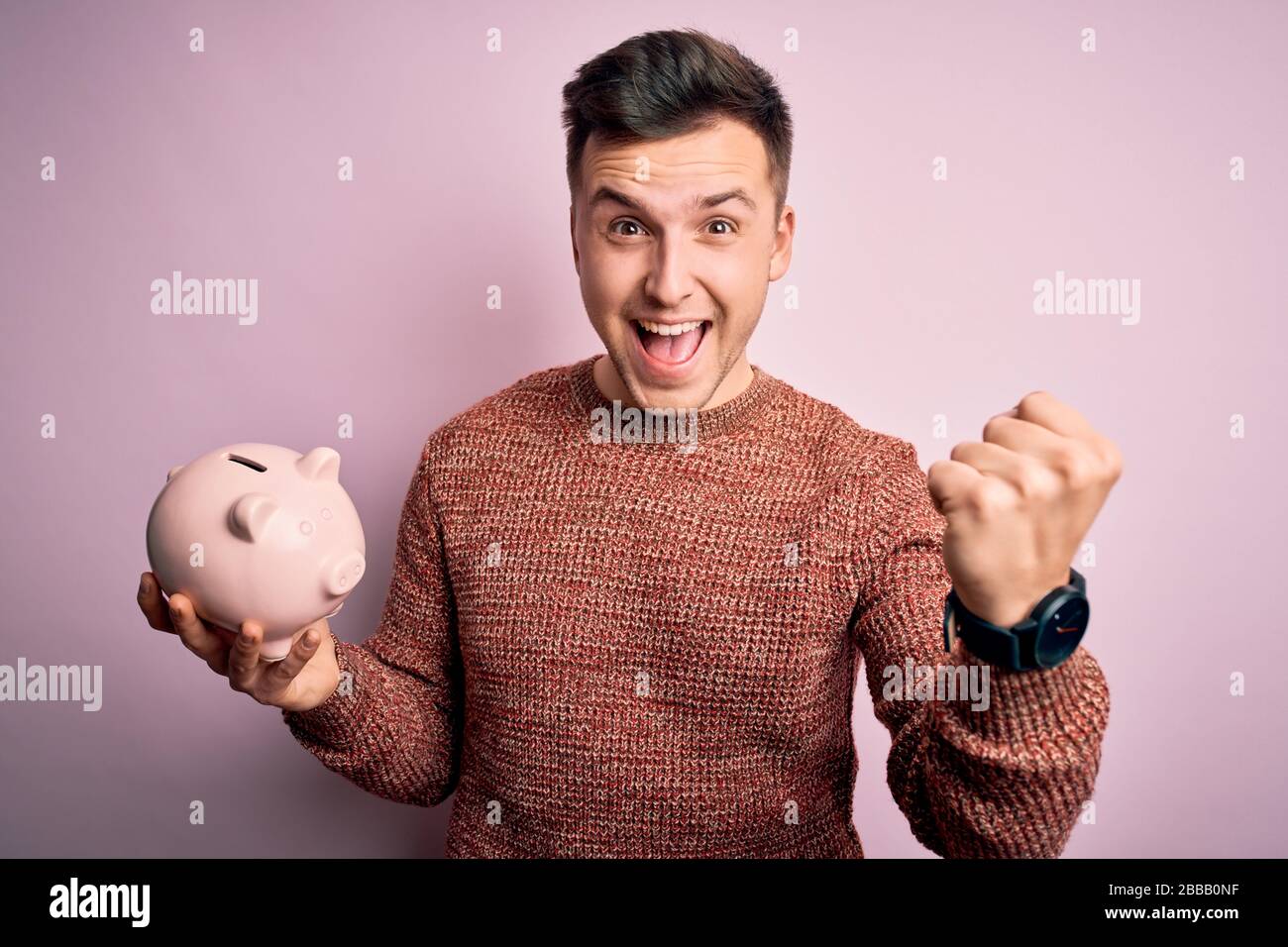 Young handsome caucasian man holding piggy bank for savings over pink ...