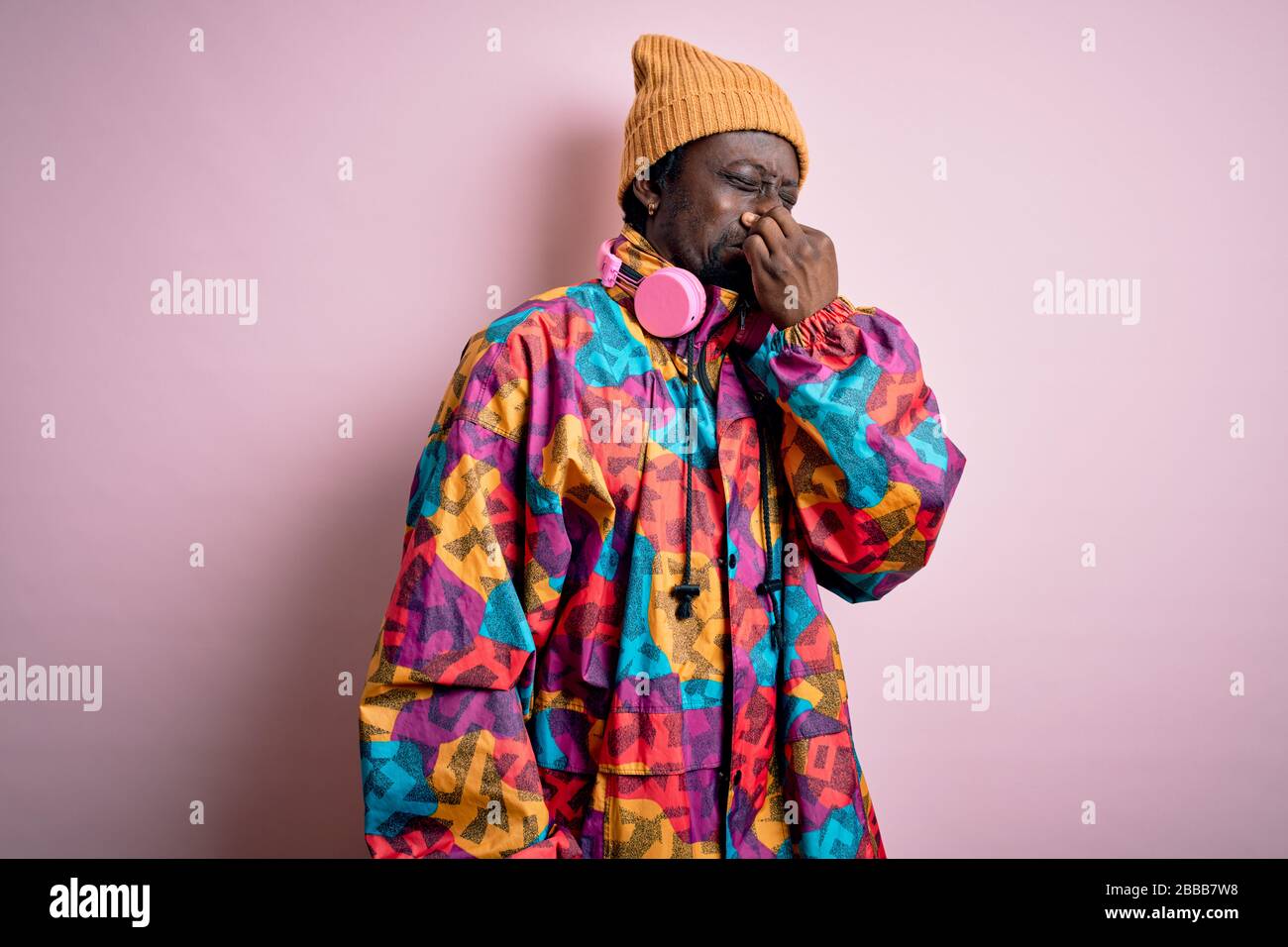 Young handsome african american man wearing colorful coat and cap over pink background smelling something stinky and disgusting, intolerable smell, ho Stock Photo