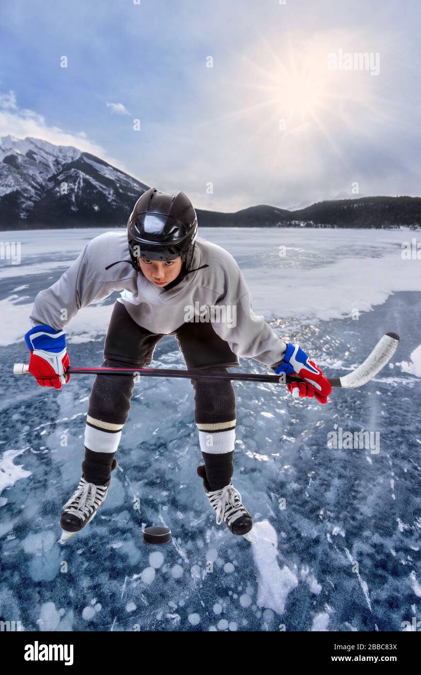 Junior Ice hockey player wearing helmet, jersey and holding hockey stick over puck in outdoor action surrounded by mountains in the Canadian Rockies. Stock Photo