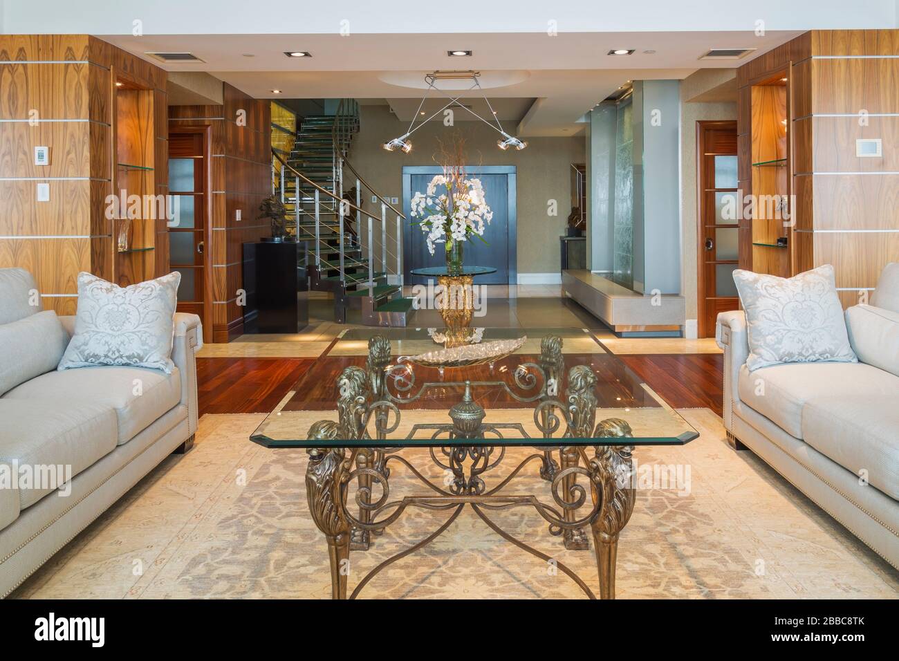 Glass top coffee table and sofas in living room with rug and exotic wood plank floor plus glass vase with white flowers on top of Chinese lion figure pedestal base with a glass top plus steel and tempered glass twisting staircase and glass wall waterfall in entryway inside a modern luxurious multistory penthouse condominium unit, Old Montreal, Quebec, Canada. This image is property released for publication in calendars and editorial use. EUPR0359 Stock Photo