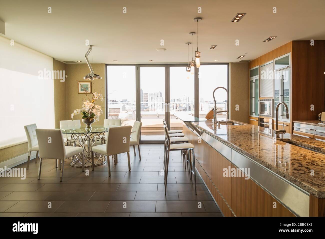 Round glass dining table with white leather chairs, wood and glass cabinets and island with grey nuanced granite countertop and white leather high back chairs in kitchen with grey marble tile floor inside a modern luxurious multistory penthouse condominium unit, Old Montreal, Quebec, Canada. This image is property released for publication in calendars and editorial use. EUPR0359 Stock Photo