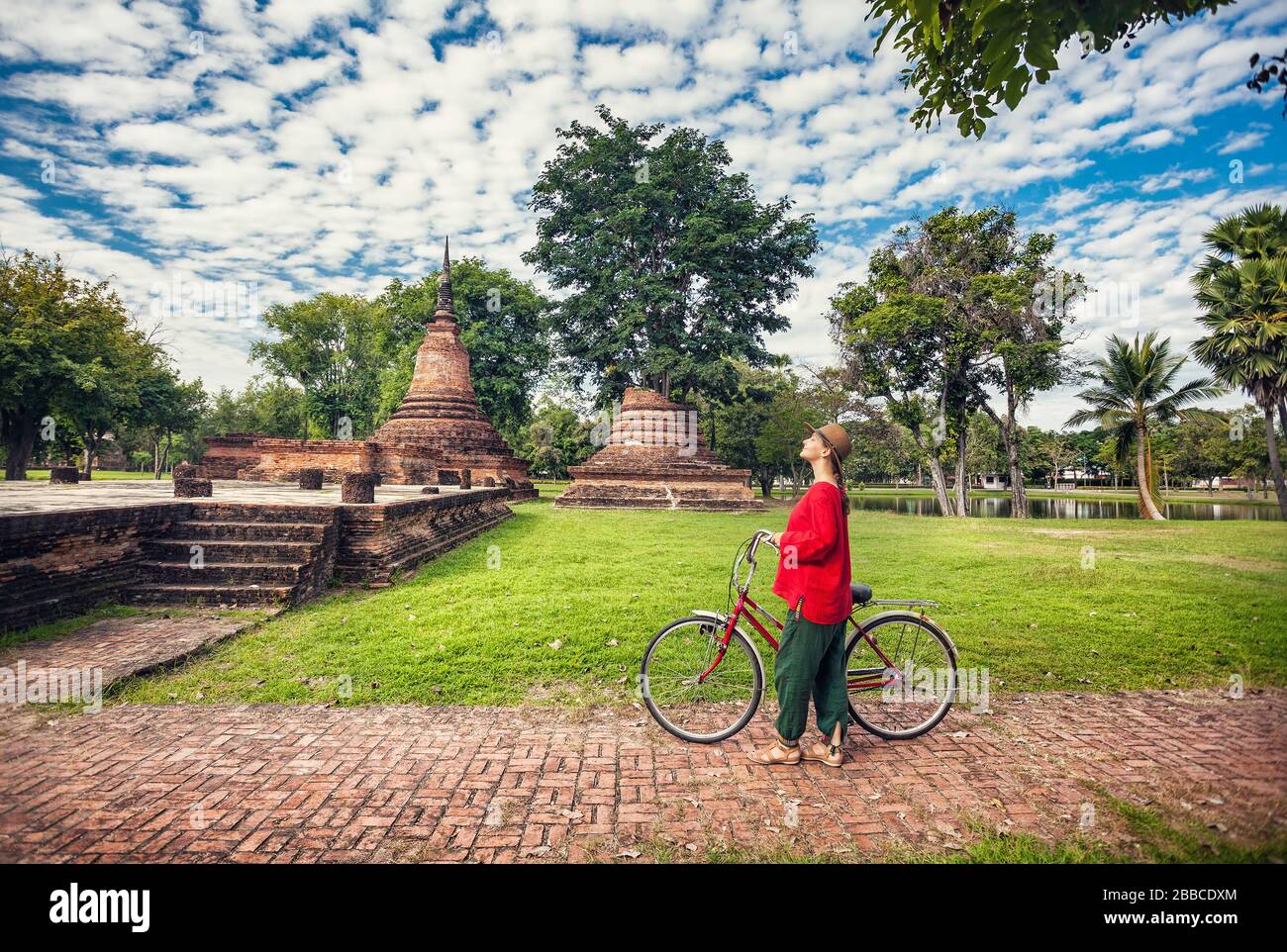 Woman in red shirt with bicycle looking at old ruined Buddhist temple in Sukhothai historical park, Thailand Stock Photo