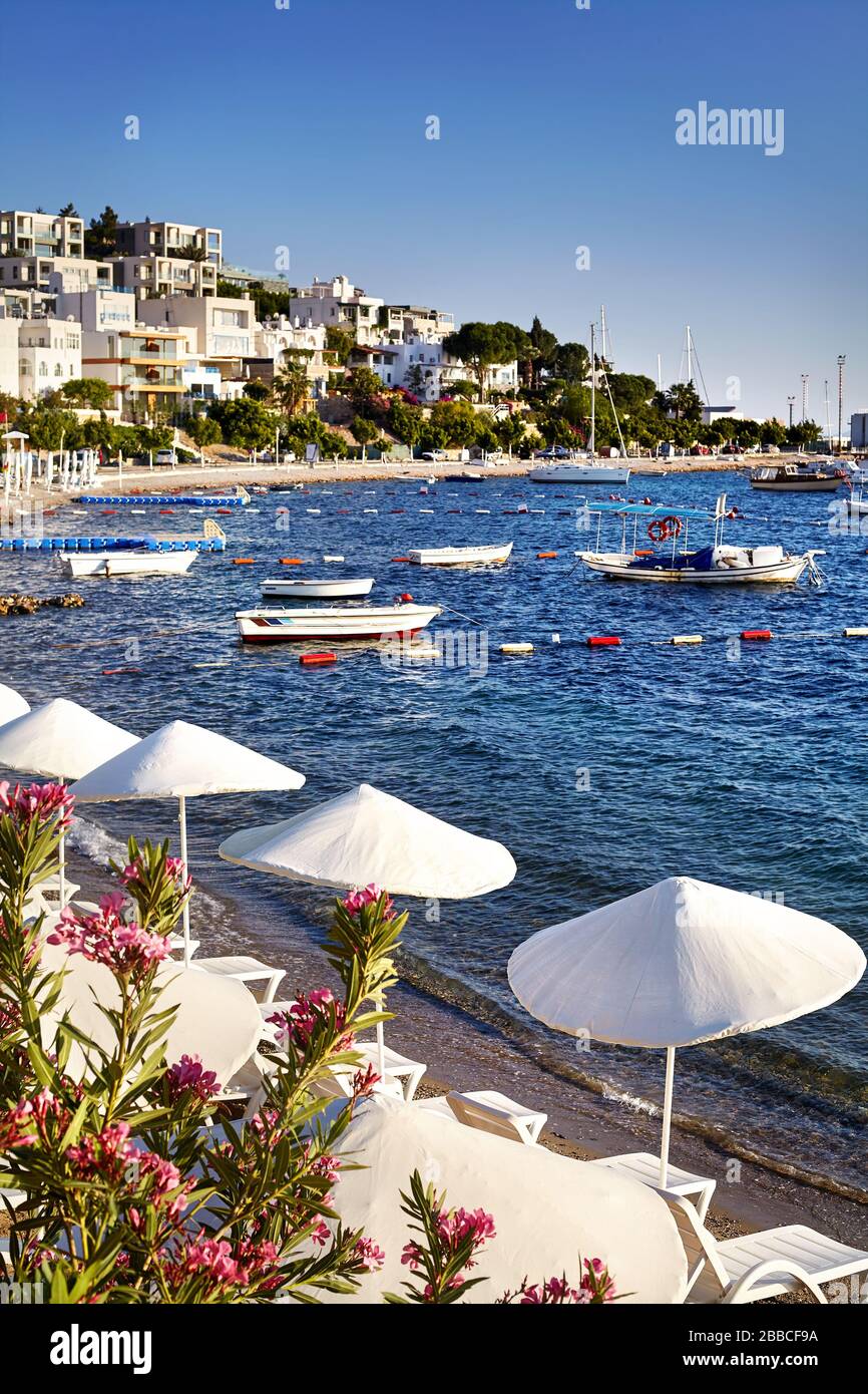 White Umbrellas and sunbeds near lagoon with boats on the beach in Bodrum, Turkey Stock Photo