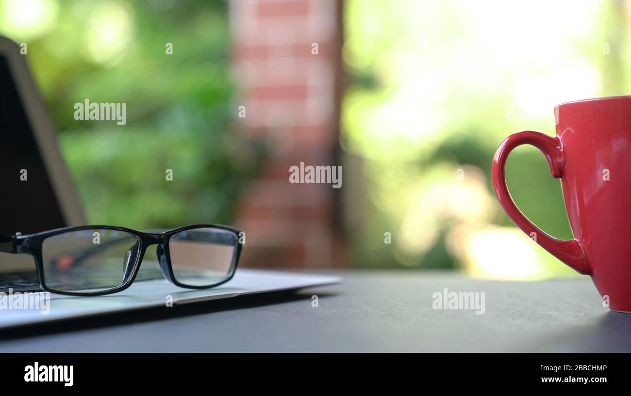Computer laptop with reading glasses and coffee in red cup with garden background. Work from home. Copy space, Stock Photo