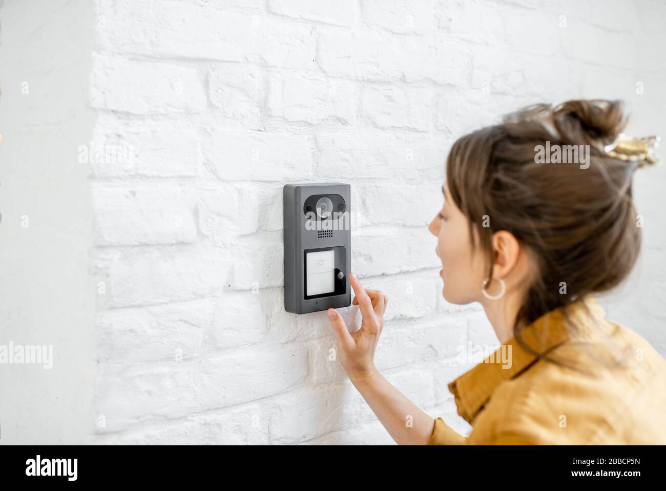 Woman rings the house intercom with a camera installed on the white brick wall Stock Photo
