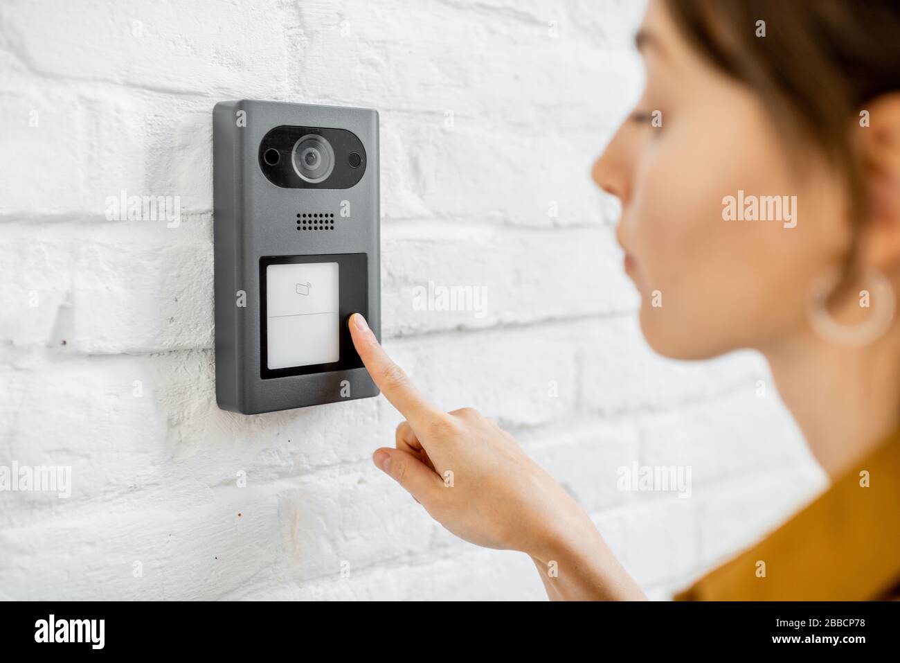 Woman rings the house intercom with a camera installed on the white brick wall Stock Photo