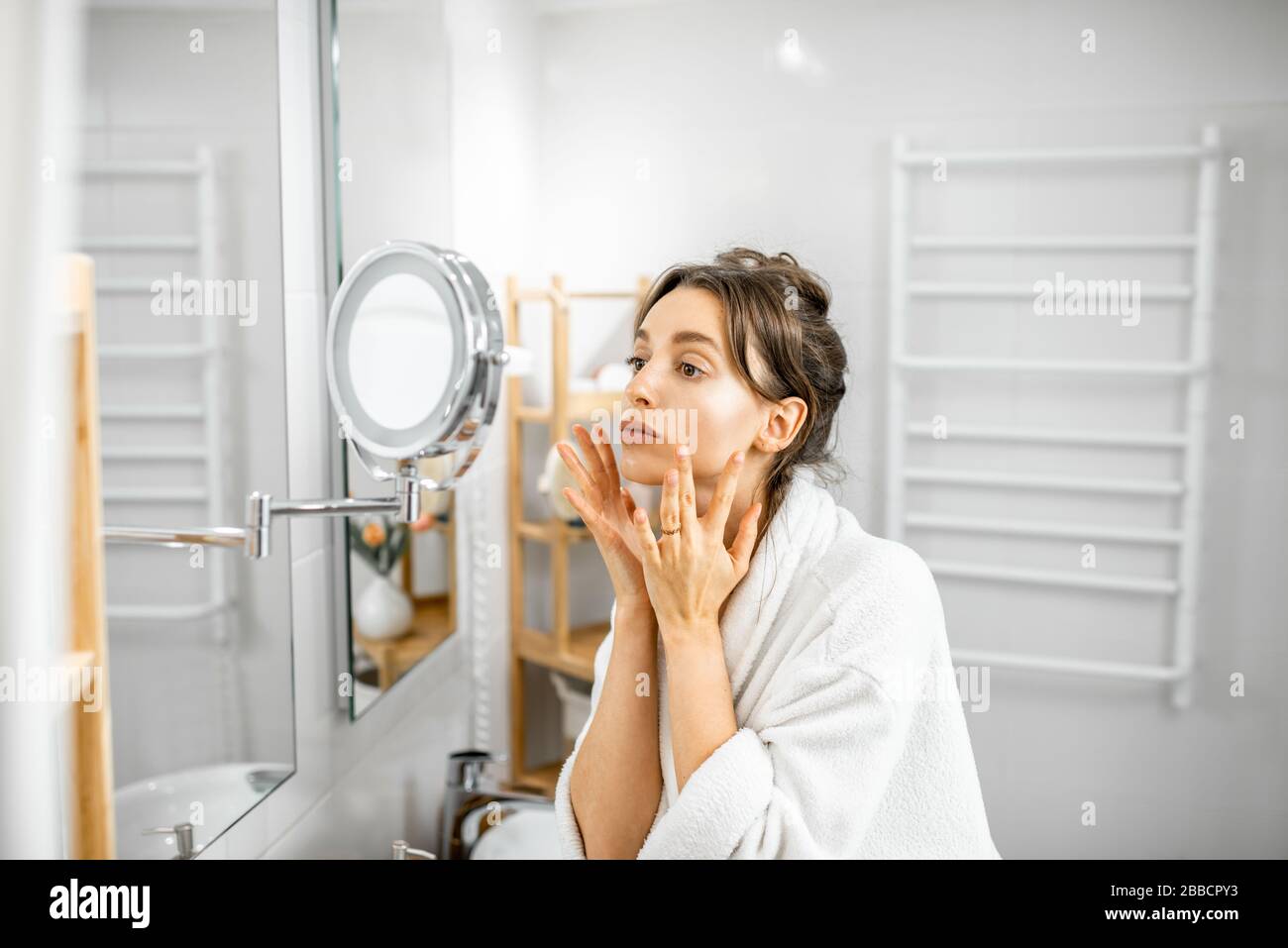 Young woman in bathrobe taking care of her skin, looking at the mirror ...
