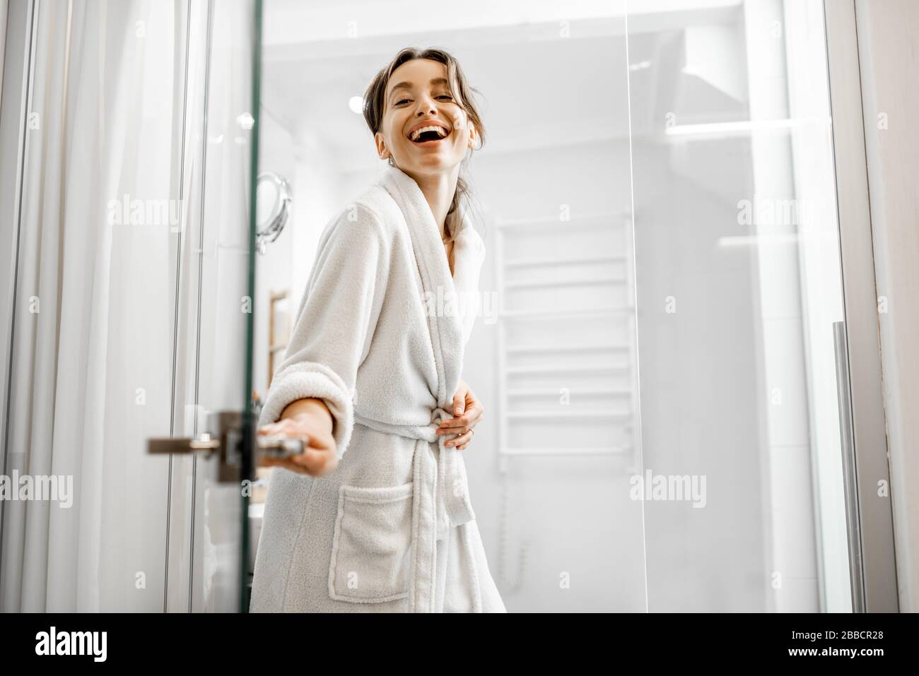 Young and joyful woman in bathrobe going to the bath, closing a glass doors at home Stock Photo