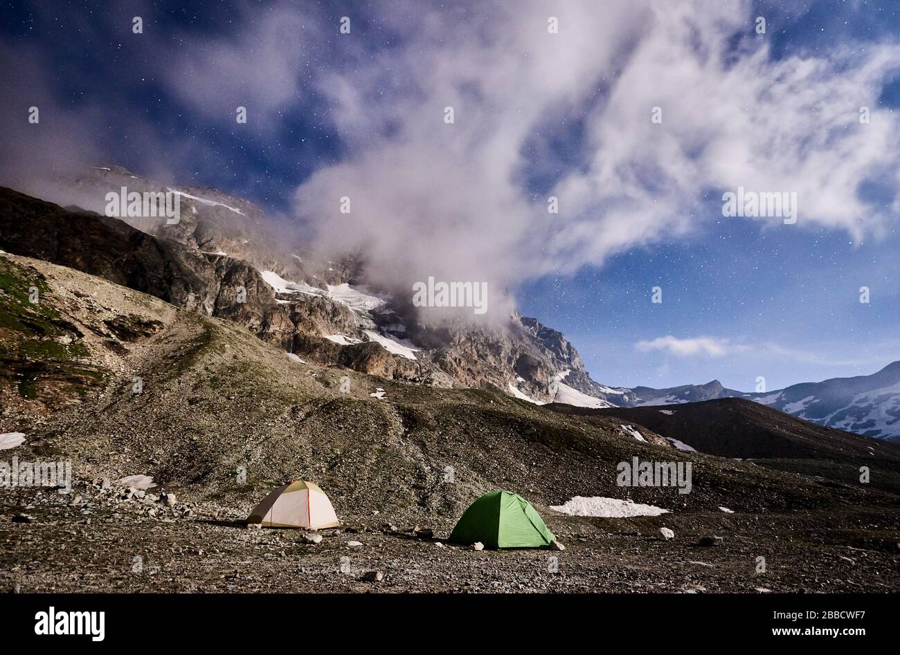 Panoramic View Of Night Camping In Mountains Under Cloudy Sky With Stars Scenery Of Tourist Tents On Rocky Hill And Mountain Peak Surrounded By Dense Mist Concept Of Travelling Hiking And Camping