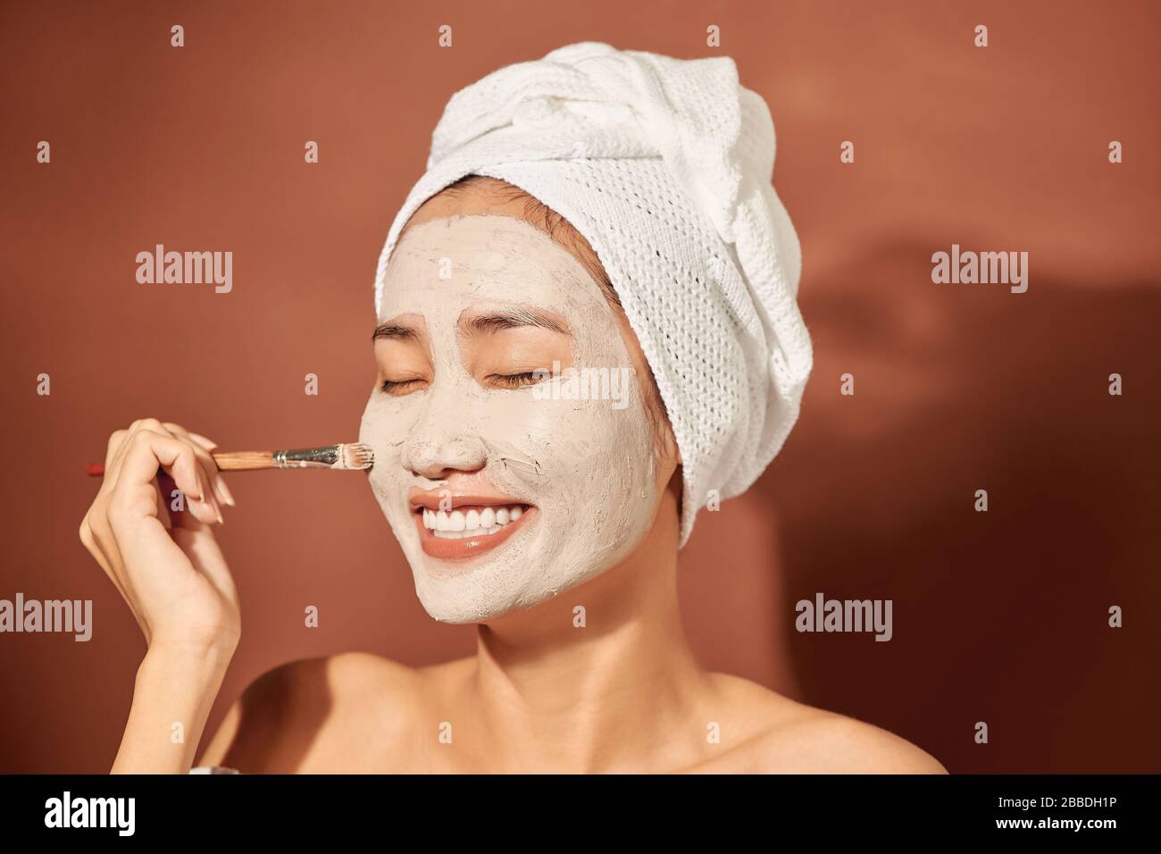 Close-up Portrait of beautiful Asian girl in spa with a towel on her head applying facial clay mask. Stock Photo