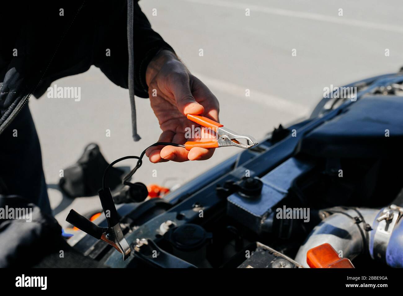 the hand connects the tongs red terminal to the battery of a car with an open hood and tuned parts of the car Stock Photo