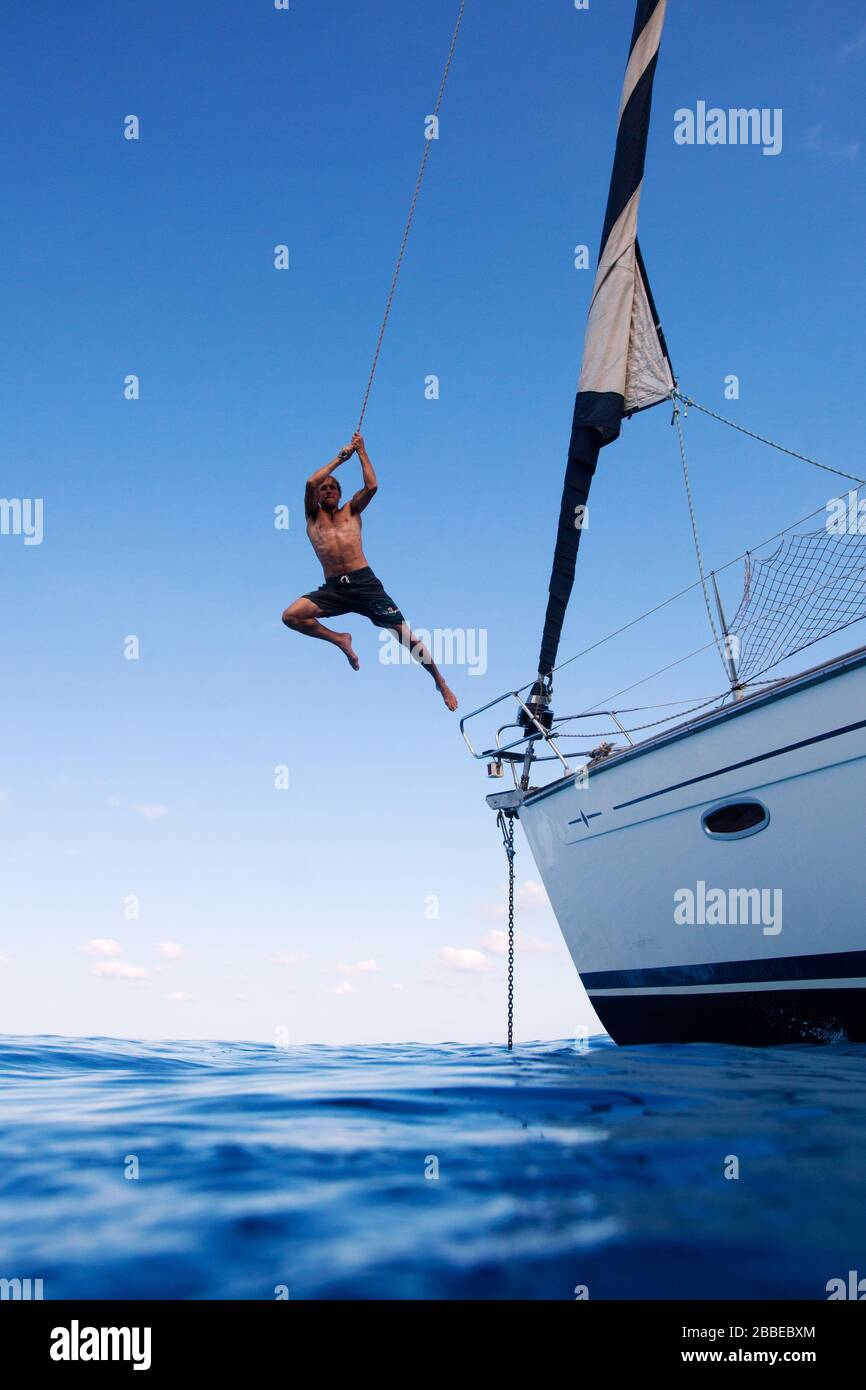 Young man swinging on a rope tied to a sailing yacht mast at Cala Goloritzé, Sardinia. Stock Photo
